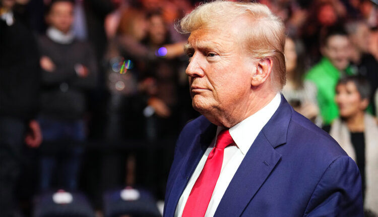 A man in a suit and red tie stands at an angle to the camera. There are many people behind him and out of focus.